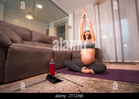 Bruna caucasica incinta in maglietta grigia che esegue esercizi di yoga. Esercizi online in classi preparatorie, benessere durante la gravidanza Foto Stock