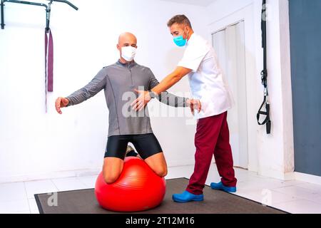 Bilanciamento client su una palla rossa assistito da fisioterapista con maschera facciale. Fisioterapia con misure protettive per la pandemia di coronavirus Foto Stock