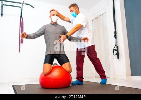 Bilanciamento client su una palla rossa assistito da fisioterapista con maschera facciale. Fisioterapia con misure protettive per la pandemia di coronavirus Foto Stock