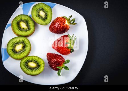 Kiwi e fragole tagliati a fette su un piatto bianco con sfondo nero Foto Stock