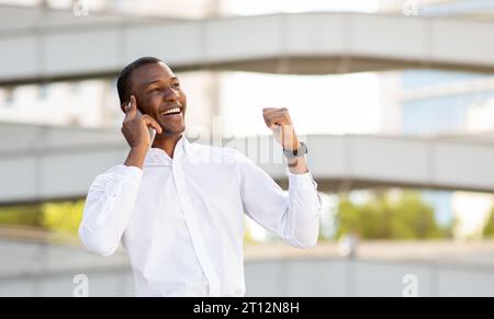 Felice uomo d'affari afroamericano che parla al cellulare all'aperto e festeggia il successo Foto Stock