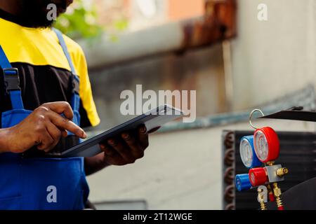 Elettricista afroamericano che esegue controlli delle perdite e altre riparazioni necessarie per evitare guasti gravi. Addetto alla manutenzione autorizzato che verifica l'aria condizionata, scrivendo i risultati sulla clipboard Foto Stock