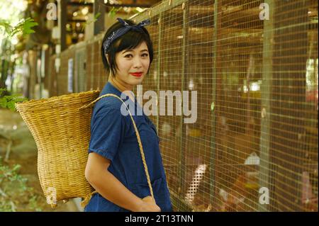 Una bella donna asiatica posa appesa a un cesto di bambù dietro il pollaio nella fattoria di polli. Foto Stock