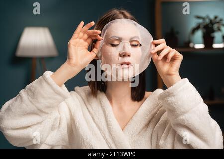 Trattamento viso e trattamenti di bellezza. Donna con un panno maschera idratante sul viso che fa procedure spa nella sua camera da letto. Copia spazio Foto Stock