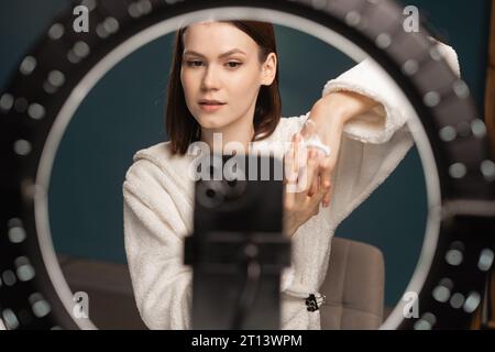 Ragazza caucasica bellezza blogger registrazione video recensione prodotto per la cura della pelle per le mani e mostrando crema alla fotocamera da condividere sui social media seduti in casa Foto Stock