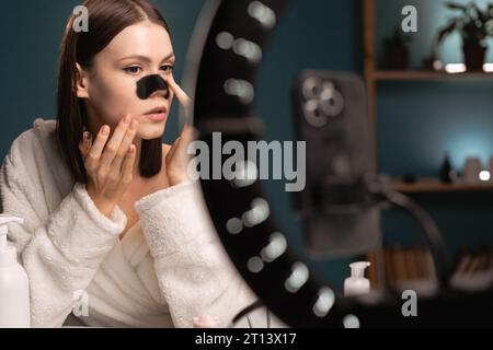 Cosmetologia. Ritratto di una bellissima blogger con maschera nera sul naso per registrare video per il suo tutorial. Trattamento di bellezza, blogging e cura della pelle. Foto Stock