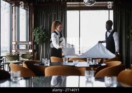 Ritratto laterale di due eleganti camerieri che ambientano i tavoli in un ristorante di lusso, con tovaglie bianche e spazio per la copia Foto Stock