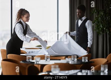 Ritratto laterale di due eleganti camerieri, tavoli con tovaglie bianche in un ristorante di lusso che si prepara all'apertura Foto Stock