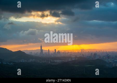 Al crepuscolo, il sole sprofonda tra le nuvole e splende sulla città di Taipei. Sagome di edifici cittadini. Cielo arancione e nuvole scure. Foto Stock