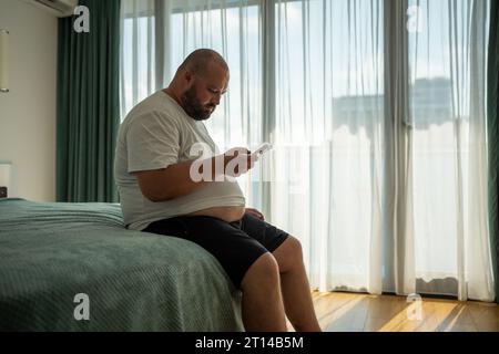 Ragazzo in sovrappeso appeso allo smartphone sul resort seduto nel letto della camera d'albergo in vacanza. Foto Stock