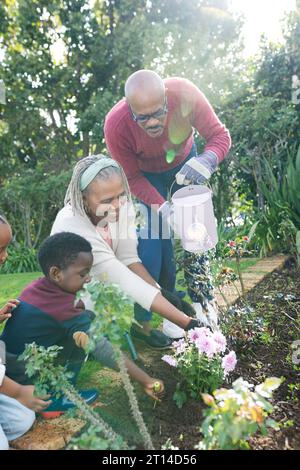 Felici nonni e nipoti afroamericani che annaffiano le piante in un giardino soleggiato Foto Stock