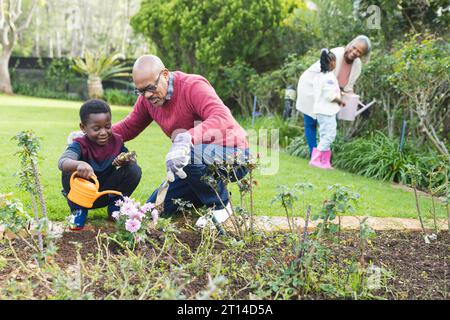 Felici nonni e nipoti afroamericani che annaffiano le piante in un giardino soleggiato Foto Stock