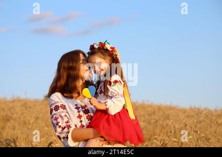 Mamma e figlia ucraine sorridenti in vyshyvanka (camicie ricamate) con un cuore giallo-blu su uno sfondo di campi di grano e cielo. Ucraina Foto Stock