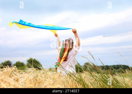 Bandiera gialla e blu dell'Ucraina nelle mani di una bella ragazza con una camicia ricamata e una corona con nastri. Bambino in un campo di grano. Independen Foto Stock