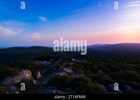 Vista al tramonto del sole che tramonta su una catena montuosa con rocce e cespugli. Splendido cielo con tonalità blu, viola e arancione. Alto da Groba - Baiona - Foto Stock