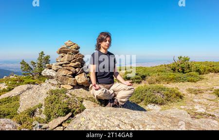 Donna caucasica di mezza età seduta accanto a una pietra miliare in posizione facile per un esercizio di meditazione in mezzo alla natura. Foto Stock