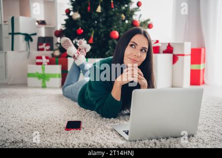 Foto di un pullover verde natalizio che indossa una ragazza premurosa e che sceglie i regali un dispositivo moderno in casa Foto Stock