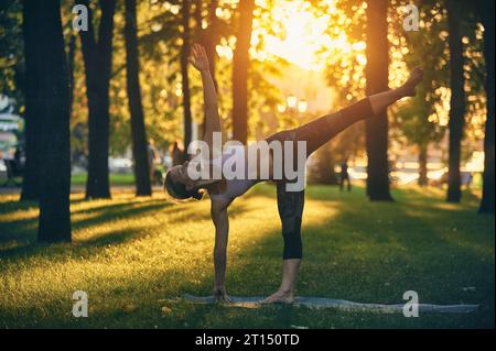 La bella donna yogini sportiva pratica yoga asana Ardha Chandrasana - mezza luna posa nel parco al tramonto Foto Stock