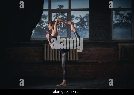 La bella donna yogi sportiva pratica yoga asana Natarajasana - posa del Signore della danza nella sala buia. Foto Stock