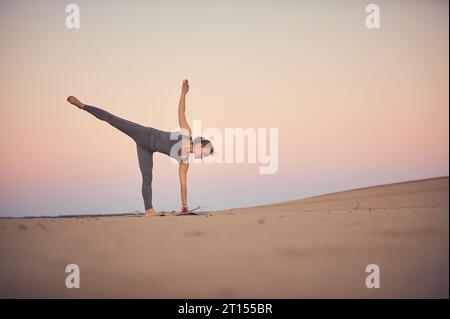 La bella giovane donna pratica yoga asana Ardha Chandrasana - mezza luna posa nel deserto al tramonto. Foto Stock