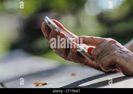 Su un parco di legno aspro, le dita di un anziano lottano con un telefono. La tecnologia di oggi, essendo piccola e sensibile, spinge gli anziani verso l'isolamento sociale Foto Stock