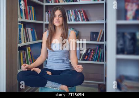 Bella donna pratica yoga asana Ardha Padmasana - mezza posa di loto nella biblioteca. Foto Stock