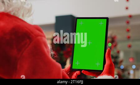 Attore del centro commerciale decorato per Natale che impersona Babbo Natale con un tablet con tasti cromatici, che si prende una pausa dal lavoro per guardare i video sullo schermo del mockup mentre i clienti girano intorno, da vicino Foto Stock