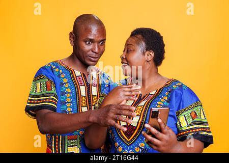 Sorridente coppia di uomini e donne adulti che discutono e leggono notizie sullo smartphone. Moglie e marito neri che indossano abiti etnici che usano il telefono cellulare insieme e parlano Foto Stock