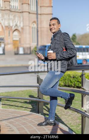 Un uomo latino che tiene una tazza di caffè usa e getta appoggiato su una ringhiera in un parco pubblico. Foto Stock