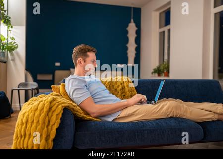 Uomo seduto sul divano con un computer portatile sul grembo per studiare, rilassarsi, rinfrescarsi, navigare in Internet, controllare le notizie Foto Stock