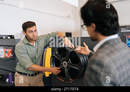 Un ritratto di un venditore professionista in tuta da lavoro che parla delle caratteristiche dei cerchi in lega al cliente maschio è venuto a vedere Foto Stock