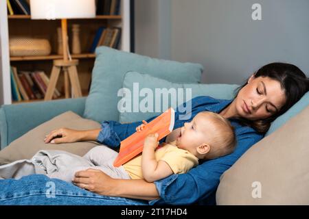 La giovane madre si addormentò dopo aver letto al suo bambino un libro di favole. Mamma stanca che dorme con suo figlio in grembo ha cercato di leggere a suo figlio in t Foto Stock