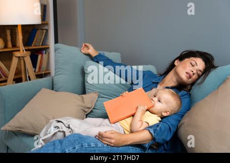 La giovane madre si addormentò dopo aver letto al suo bambino un libro di favole. Mamma stanca che dorme con suo figlio in grembo ha cercato di leggere a suo figlio in t Foto Stock