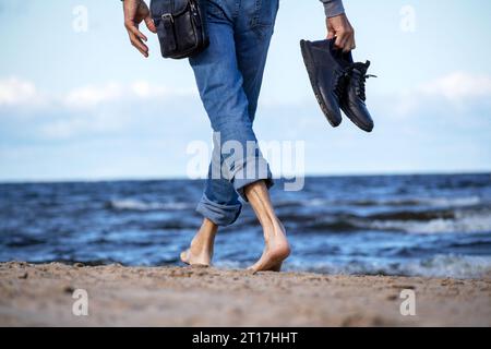 Sulla spiaggia di colore blu si erge un uomo con pantaloni in denim con scarpe nere in mano Foto Stock