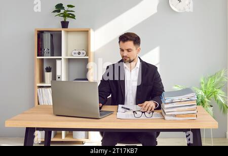 Uomo d'affari concentrato che lavora a casa con computer portatile e documenti sulla scrivania Foto Stock