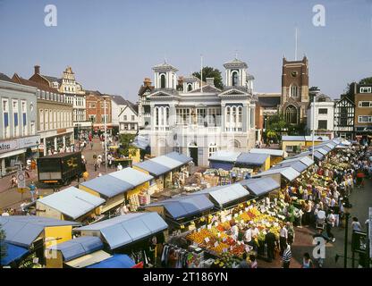 Vista aerea dell'archivio storico del 1991 nell'antico e affollato mercato di Kingston, una città nel Royal Borough di Kingston sul Tamigi, nel sud-ovest di Londra, con i commercianti che servono gli acquirenti da sotto le tettoie, bancarelle coperte e torri gemelle in posizione centrale di edificio del mercato del vecchio municipio accanto ai nomi commerciali della chiesa di tutti i Santi e della torre elencati di grado i del XII secolo include Woolworths & Curry's Foto Stock