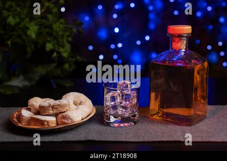 Pasta frolla, ciambelle di vino e pasticceria sfoglia su un piatto di legno e accanto a un bicchiere con ghiaccio e una bottiglia di liquore, con uno sfondo di luci blu Foto Stock