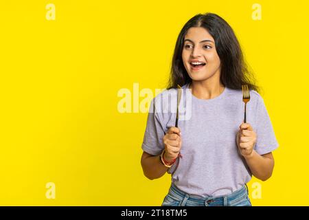 Pronti a mangiare. Una donna indiana affamata che aspetta di servire piatti per la cena con un coltello da forchetta per posate senza irrequietezza apprezzerà il delizioso pasto del ristorante. Ragazza eccitata su sfondo giallo Foto Stock