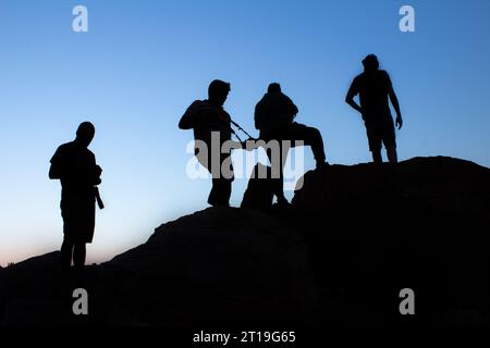 Silhouette di un gruppo di fotografi maschi in piedi o che scattano fotografie in cima alla montagna al tramonto serale con cielo blu sullo sfondo Foto Stock