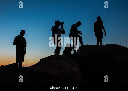 Silhouette di un gruppo di fotografi maschi in piedi o che scattano fotografie in cima alla montagna al tramonto serale con cielo blu sullo sfondo Foto Stock