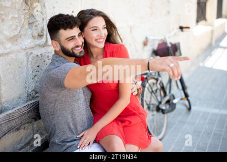 Giovane coppia sorridente e felice. Seduto su una panchina accanto a una bicicletta. Foto Stock