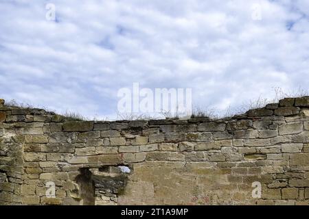 Un vecchio muro di un antico edificio in pietra distrutto dal tempo con erba secca sullo sfondo di un cielo nuvoloso. Copia spazio. Messa a fuoco selettiva. Foto Stock