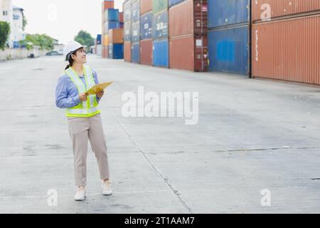 La donna asiatica lavoratrice di banchina felice controlla il carico dei container nei cantieri navali. Dipendente responsabile del personale marittimo e di trasporto. carico di carico per la spedizione da parte dell'operatore Foto Stock