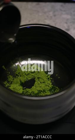 Il barista versa in polvere di tè verde matcha biologico in un recipiente con un cucchiaio di legno. Preparazione del tè verde Matcha in una ciotola nera su un tavolo di legno Foto Stock