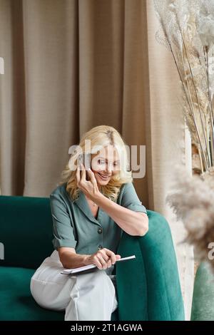 donna d'affari sorridente di mezza età con i capelli biondi che parla sullo smartphone e tiene in mano il notebook Foto Stock