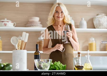 bella donna di mezza età con capelli biondi che regge un bicchiere con vino rosso in cucina moderna Foto Stock