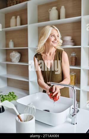 allegra donna di mezza età che lava pomodori ciliegini freschi e maturi e sorride in una cucina moderna Foto Stock