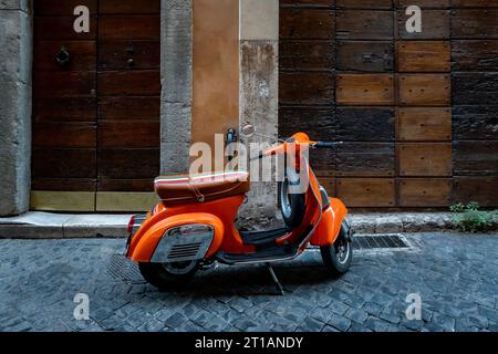 Scooter arancione, vespa parcheggiata accanto al muro con porte in legno sulla strada asfaltata vuota Foto Stock