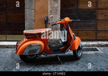 Scooter arancione, vespa parcheggiata accanto al muro con porte in legno sulla strada asfaltata vuota Foto Stock