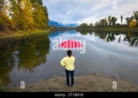 Una donna con un ombrello rosso si trova sulle rive del tranquillo fiume Kootenai vicino a Bonners Ferry, Idaho. Foto Stock